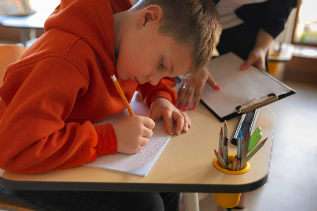 Chłopiec w pomarańczowej bluzie z kapturem siedzi przy biurku i z dużym skupieniem pisze ołówkiem w zeszycie. Obok niego stoi dorosła osoba trzymająca podkładkę z dokumentami