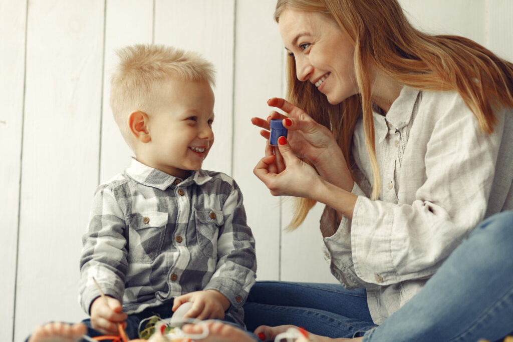 Kobieta o blond włosach i mały chłopiec w koszuli w kratę siedzą naprzeciwko siebie, śmiejąc się. Terapeuta pokazuje dziecku mały niebieski przedmiot, angażując je w interakcję. Scena emanuje bliskością i wsparciem emocjonalnym.