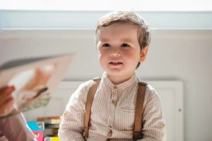 Portret małego chłopca z zaciekawioną miną, siedzącego przed osobą trzymającą otwartą książeczkę z ilustracjami. Jasne oświetlenie i bliski plan na twarz dziecka podkreślają znaczenie interakcji i komunikacji we wczesnym dzieciństwie.